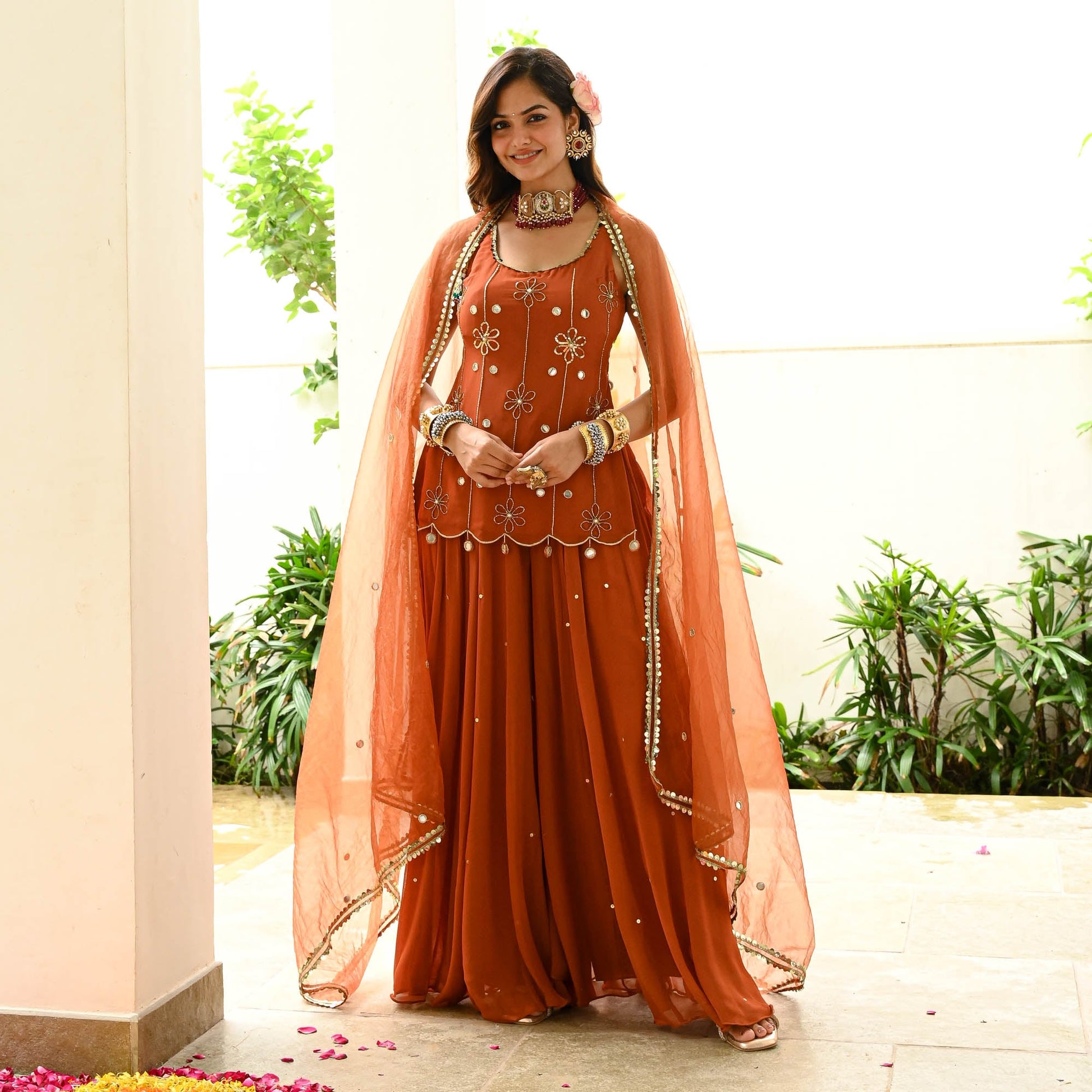 Woman in an orange traditional outfit with a dupatta, standing indoors with plants in the background.